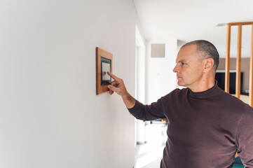 A man in a brown turtleneck shirt interacting with a digital control panel mounted on a wall. He is...