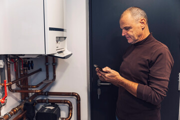 A man adjusting a digital timer on a wall-mounted heating control panel. The panel features multiple dials and buttons, with copper pipes visible in the background.