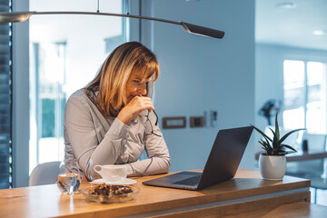 Obraz premium A woman sitting at a wooden table, looking at her laptop with a thoughtful expression. She has a cup of coffee and a glass of water beside her, with a small plant in the background. The setting is bri