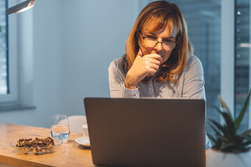 A woman with glasses is sitting at a wooden table, focused on her laptop. She has a thoughtful...