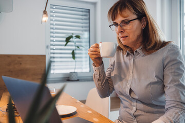 A woman in a striped shirt sitting at a wooden table, holding a cup while looking at a laptop. She appears focused and is in a modern, well-lit room with plants in the background.