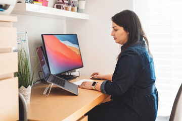 A woman working at a desk with a computer and a laptop. She is focused on her work, sitting in a modern home office environment. The desk is tidy with a few plants and office supplies.