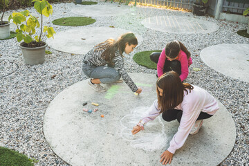 Three children drawing with chalk on a concrete surface in a garden. The setting features gravel...