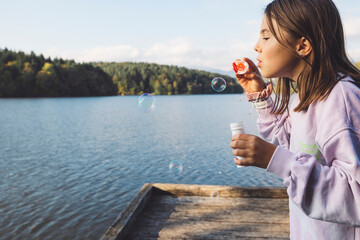A young girl blowing bubbles by a serene lake, surrounded by trees. She wears a light purple sweater and has a joyful expression. The bubbles float in the air, reflecting sunlight.