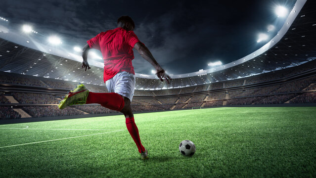 Dynamic image of man, soccer player in red uniform, kicking ball on well lit stadium field at night. Energy and focus. 3D render of stadium. Concept of sport, competition, tournament, games, event