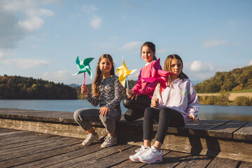 Fototapeta premium Three children sitting on a wooden dock by a lake, holding colorful pinwheels. They are smiling and enjoying a sunny day with a scenic background of trees and blue sky.
