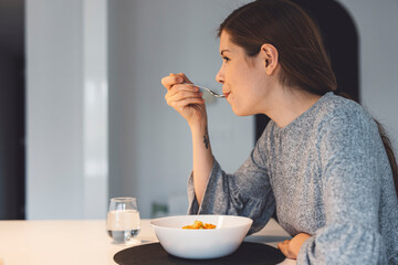 A young woman sitting at a dining table, enjoying a bowl of salad. She has long brown hair tied...