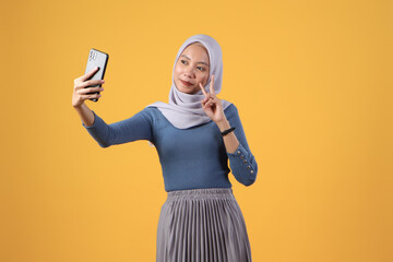 happy asian indonesian muslim woman holding smart phone giving victory finger gesture on isolated yellow background