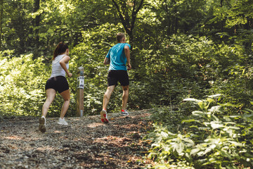 A man and a woman running on a gravel path through a lush green forest. The woman is in a white tank top and shorts, while the man wears a blue shirt and black shorts. Sunlight filters through the tre