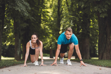 A man and woman in athletic attire are positioned in a starting stance on a path surrounded by...