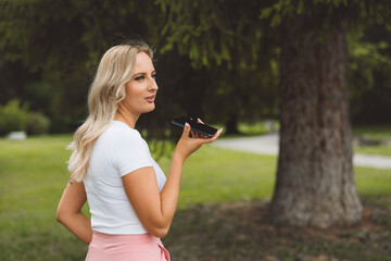 A young woman with long blonde hair is standing outdoors, holding a smartphone to her ear. She is wearing a white top and pink pants, looking thoughtfully into the distance. The background features gr