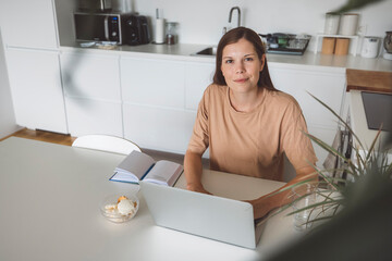 A woman sitting at a table with a laptop, looking thoughtfully at the screen. She has long brown hair and is wearing a beige shirt. A notebook and a small bowl are on the table, with a kitchen visible