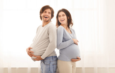 Fototapeta premium Pregnant fun. Portrait of joyful young man with big fake belly and his pregnant wife holding hands on their tummies and laughing at home, empty space