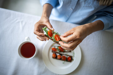 Closeup hands holding rye crisp bread with creamy vegetarian cheese tofu, cherry tomato, rucola micro greens at table with cup of tea at home. Healthy food, vegan breakfast, gluten free, morning diet.
