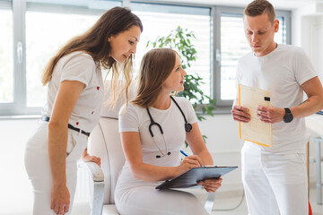 Two healthcare professionals discussing a patient's chart in a modern clinic. One is seated, reviewing documents, while the other stands beside her, pointing at the chart. Both are dressed in white at