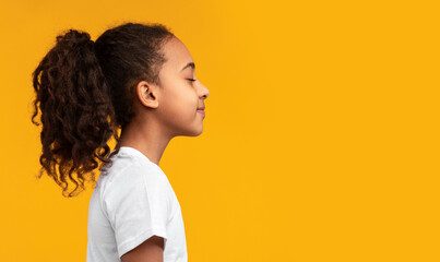 Side view profile portrait of African American girl in white t-shirt posing with closed eyes and curly ponytail hair isolated on yellow studio background wall. Empty free copy space, banner, panorama © Prostock-studio