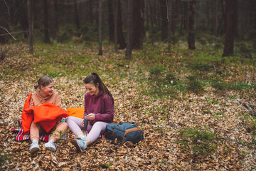Fototapeta premium Two young women sitting on a blanket in a forest having picnic, one is looking at a smartphone while the other is wearing headphones. They are surrounded by autumn leaves and trees.