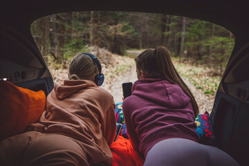 Two young women sitting in the back of a car in a forest, sharing a moment while looking at a smartphone. Free time in nature. They are wearing casual clothing and listening to music with earphones.