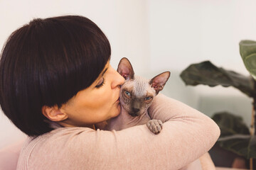 A young woman gently holds a Sphynx cat in her arms, showcasing a tender moment. The woman has long hair and wears a cozy sweater, while the cat appears calm and relaxed. The background is softly blur