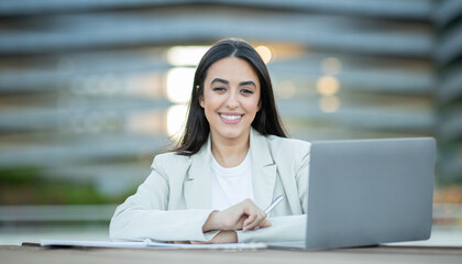 A woman sits at a table, smiling as she works on her laptop in an outdoor setting during the afternoon.