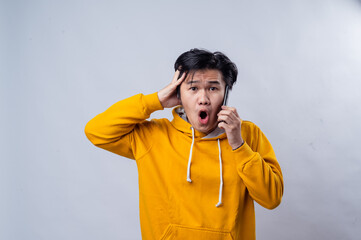 Asian man in a yellow hoodie is talking on the phone with a surprised expression. holds the phone...