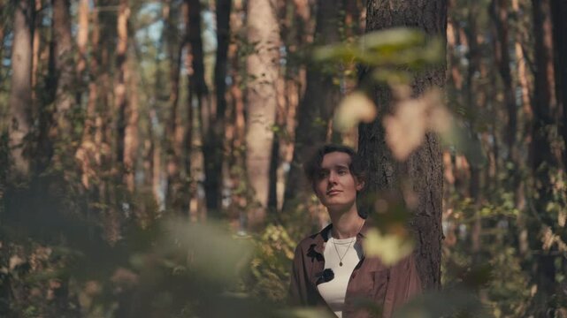 A girl of Slavic appearance in an autumn pine forest, hair in a ponytail, leaning against a pine tree, sun rays coming through the tree crowns and falling on her face, medium shot from behind the bush