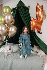 Child birthday party celebration. Happy little girl 4-5 years in stylish rustic style dress stands on bed decorated with baldachin and colorful foil balloons, smiles and looks at camera at bedroom © Andriy Medvediuk