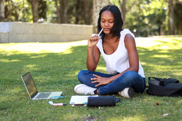 Young black woman student sitting on the grass in a park, working on her laptop and writing notes, focused on her studies, surrounded by stationery, on a sunny day