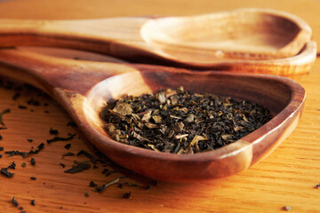 A close-up of loose leaf tea in a wooden spoon. The tea leaves are scattered around the spoon, and the wooden spoon is resting on a wooden surface