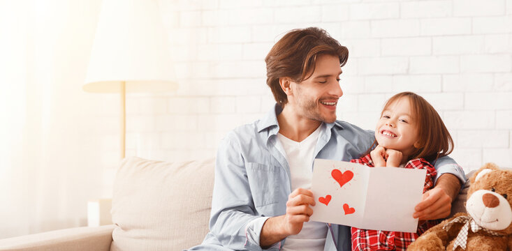 Happy man and little girl sitting on sofa at home, celebrating Father's Day, dad holding gift card and present, free space