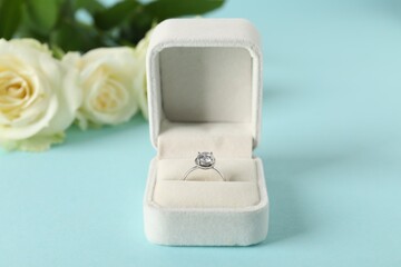 Beautiful ring with gemstone in box and flowers on light blue background, closeup