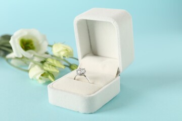 Beautiful ring with gemstone in box and flowers on light blue background, closeup
