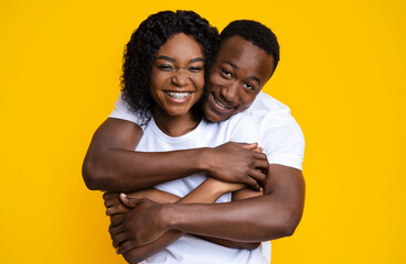 Loving black couple cuddling over yellow studio background. Cheerful young african american man and woman hugging and smiling at camera. Love, relationships, marriage concept