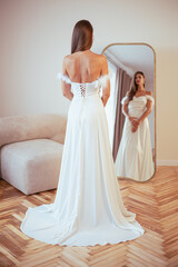 Fototapeta premium Beautiful brunette woman in a white dress. Stands in the room in front of the mirror, looks at her reflection and admires herself