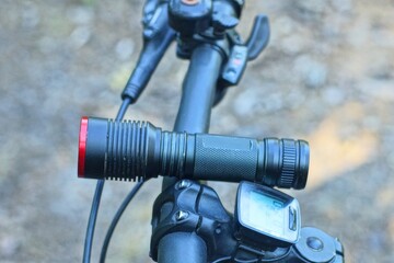 one black iron modern military glass reflector electric battery powered industrial powerful round portable flashlight lies on the handlebar of a sports mountain bike in the daytime outdoors