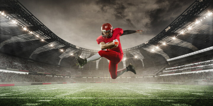 Football player in red uniform is caught mid-air as he leaps high, holding ball tightly, with stadium in background. 3D render of stadium. Concept of sport, tournament, competition, game event