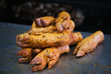 Seasoned pork hooves on a dark metallic surface, prepared for smoking with a blurred background suggesting a rustic setting