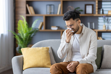 Fototapeta premium A young Indian man sits at home on the couch and wipes his nose with a tissue from a runny nose, suffering from an illness