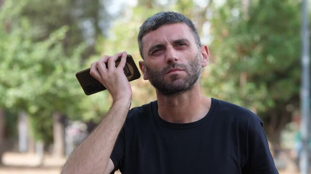 Medium shot of a man listening carefully to a voice message on his smartphone in a park. His focus on the device is evident while surrounded by nature
