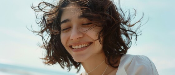 Naklejka premium A young woman with windblown hair laughs joyfully, radiating happiness and carefree energy by the seaside on a breezy day.