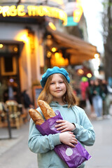Portrait of pretty young french teenager girl in blue beret holding baguettes against the backdrop of the evening lights of a cafe or bakery on a narrow Parisian street
