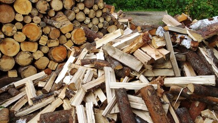a pile of chopped firewood. In the background, there is a neatly stacked pile of round logs, while in the foreground, there is a more scattered pile of split wood pieces