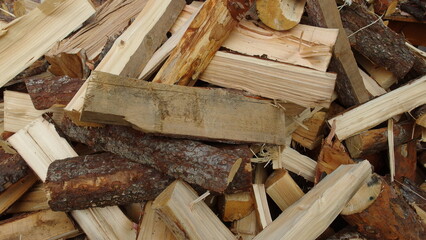  pile of chopped firewood. The pieces of wood vary in size and shape, with some having bark still attached and others being more smooth and split. The wood appears to be a mix of lighter and darker 