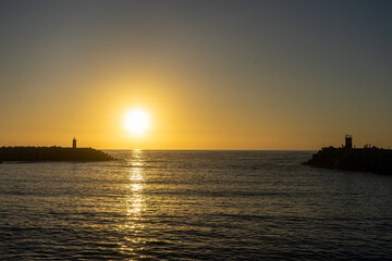 A breathtaking sunset at Nazaré, Portugal, with the sun dipping below the horizon, framed perfectly between two lighthouses at the entrance of the marina, casting golden reflections on the sea.