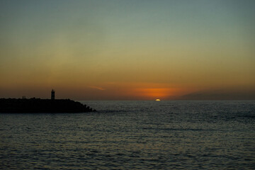 Obraz premium A breathtaking sunset at Nazaré, Portugal, with the sun dipping below the horizon, framed perfectly between two lighthouses at the entrance of the marina, casting golden reflections on the sea.