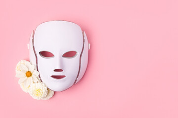 Cosmetic LED facial mask regenerative treatment on pink background with flowers dahlias. Health and beauty of the face.