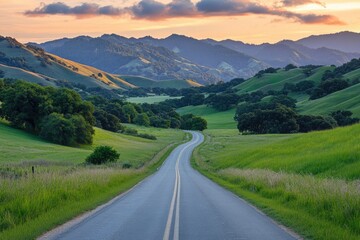 Fototapeta premium A scenic road winds through lush green fields, framed by majestic mountains under a colorful, sunset sky.