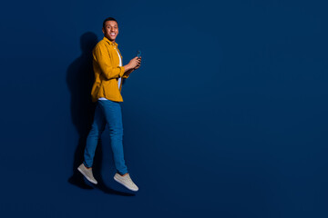 Photo of young guy wear jeans and yellow shirt jumping hold smartphone communicating isolated on dark blue color background