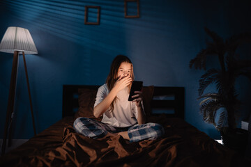 Teenage girl suffering from insomnia sitting on bed using mobile phone with shocked expression