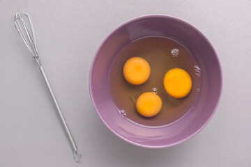 Raw eggs in purple cup with Whisk on grey background.